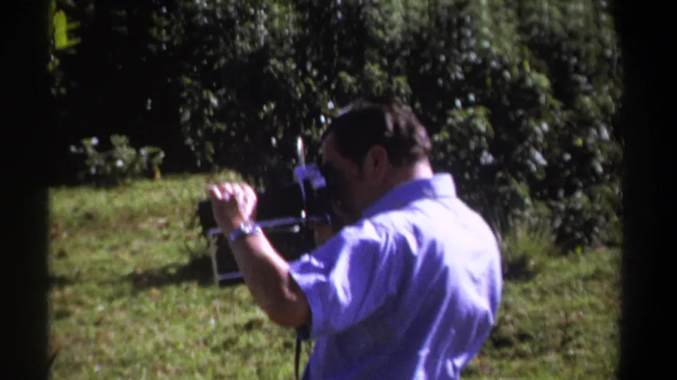 In 1969 Kenya home-movie footage, a man in a light blue shirt films outdoors with a vintage 8mm camera amid grass and trees.