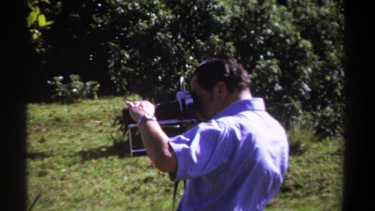 Man holds a vintage movie camera while standing on grass near dense foliage.