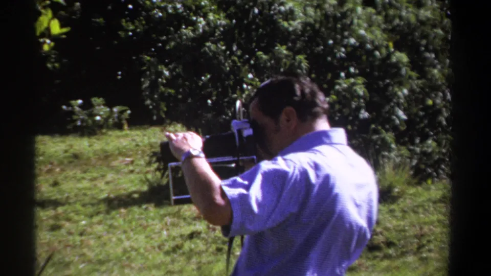 The man continues filming in a sunny Kenyan garden, holding the 8mm camera close to his face with trees behind him.