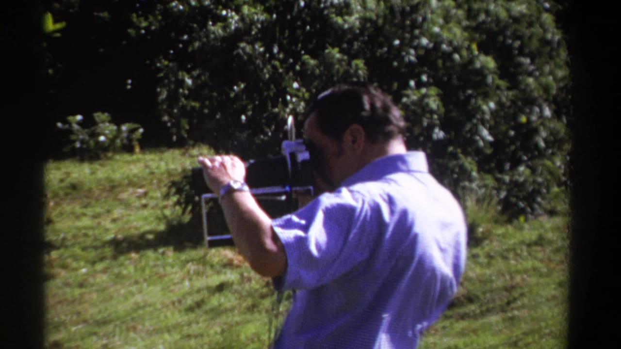 A 1969 Kenya frame shows the man steadying the 8mm camera in a green outdoor setting with soft focus and film grain.
