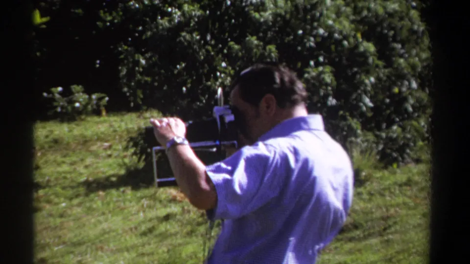 A 1969 Kenya frame shows the man steadying the 8mm camera in a green outdoor setting with soft focus and film grain.