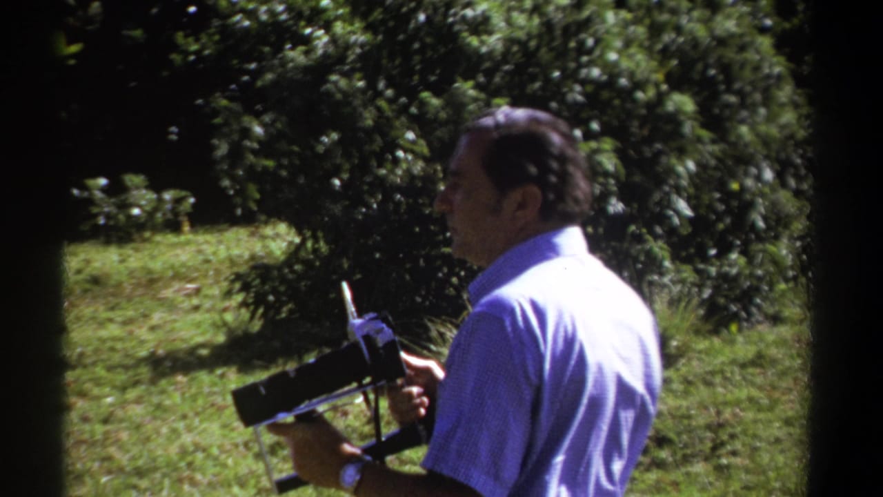 The man lowers the 8mm camera at the end of the shot, with grass, trees, and bright foliage visible in the Kenyan garden.