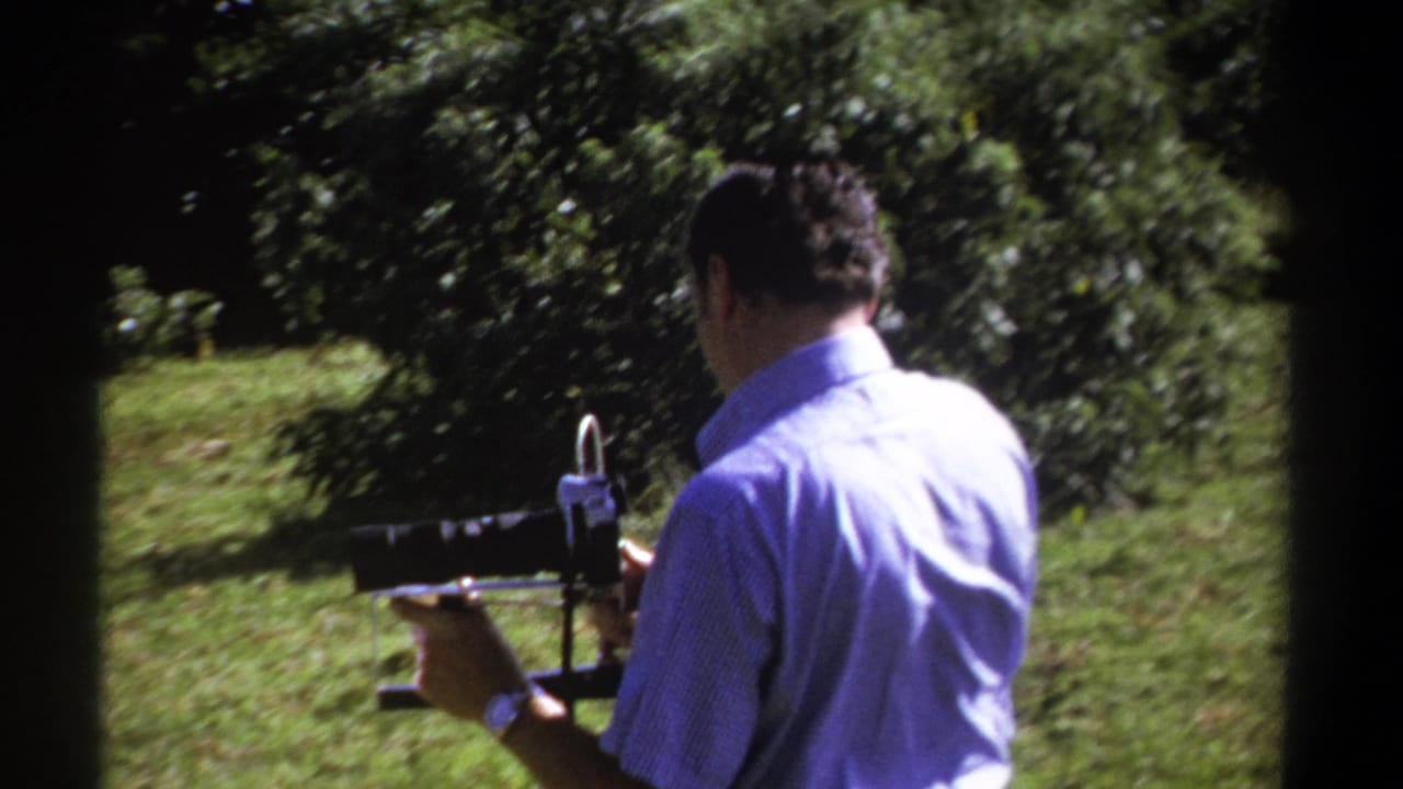 In 1969 Kenya footage, a man in a light blue short-sleeved shirt holds an 8mm camera in a sunlit green outdoor setting.