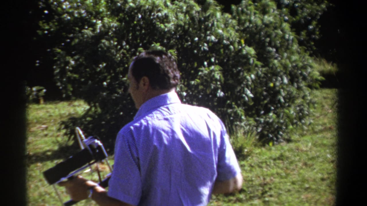 The man is viewed from behind in a Kenya 1969 home-movie frame, carrying a vintage 8mm camera beside dense green shrubs.