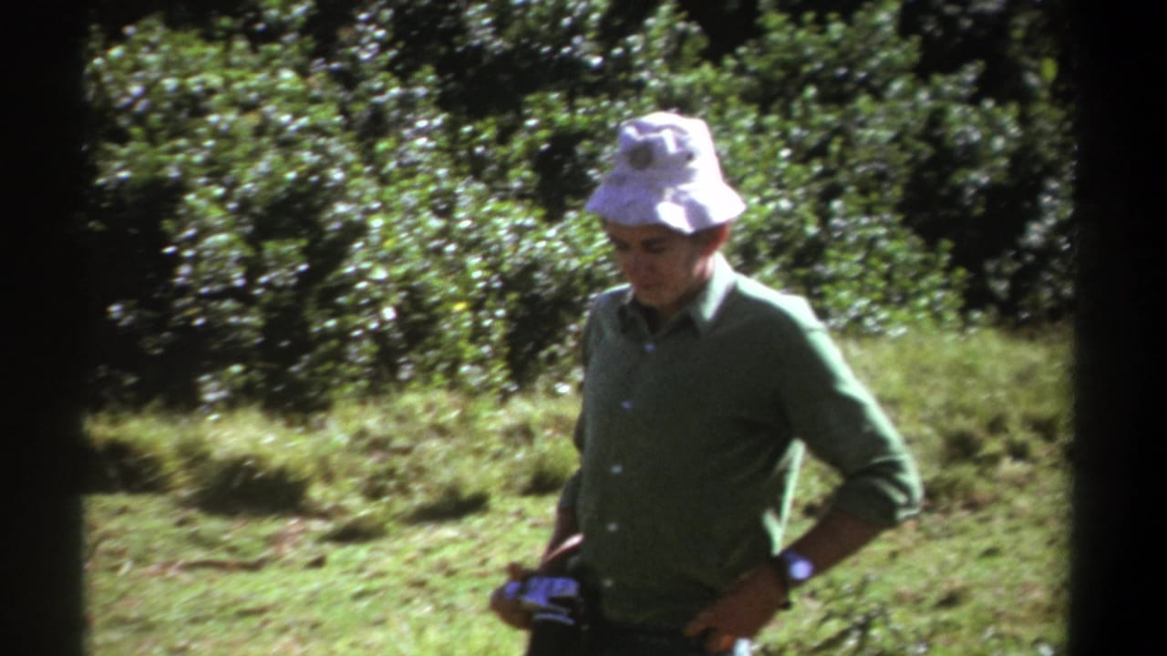 In 1969 Kenya home-movie footage, a man in a white bucket hat and green shirt stands in bright forest light holding a camera.