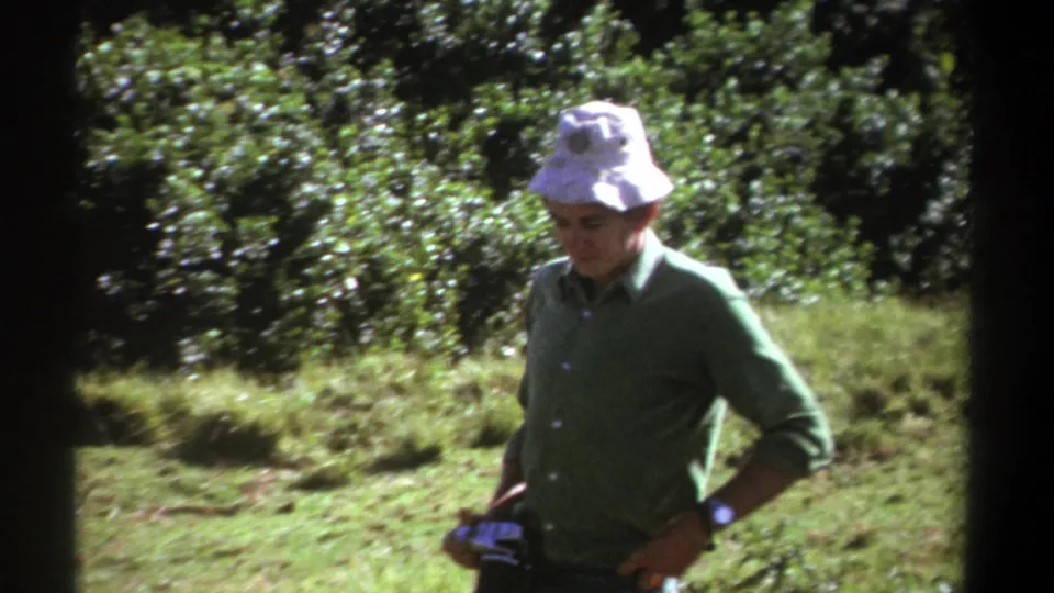 In 1969 Kenya home-movie footage, a man in a white bucket hat and green shirt stands in bright forest light holding a camera.