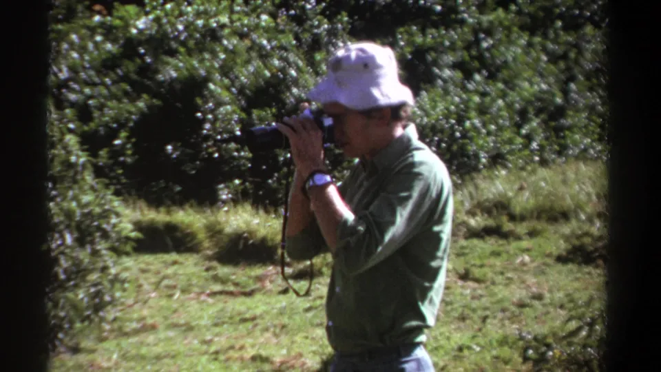 Side view of a man looking through his camera in a grassy Kenyan forest setting, captured on 8mm home-movie film.