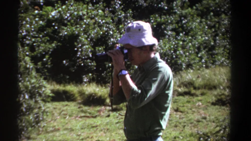 The man continues aiming his camera at forest foliage in Kenya, with grainy 8mm texture and bright outdoor exposure.