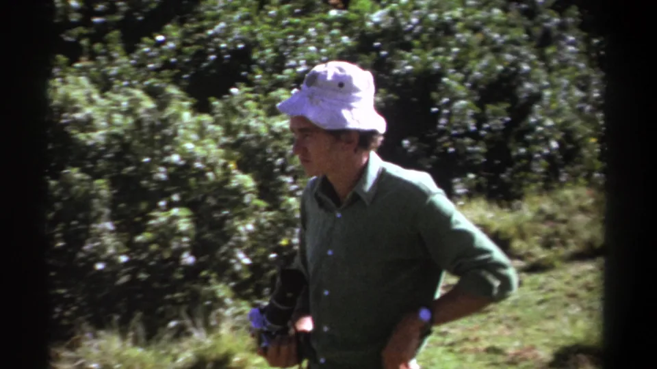 The 1969 Kenya footage shows the man paused in profile among trees, carrying a dark camera while light breaks through the foliage.