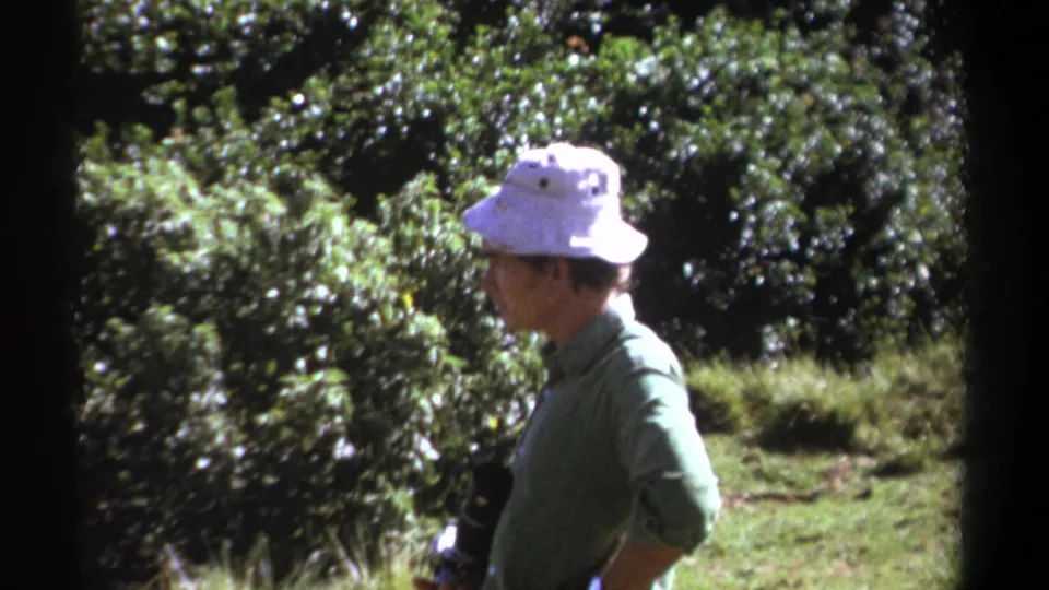 A profile view shows the adult man in a white bucket hat and green shirt moving through the Kenya forest setting in 1969 footage.