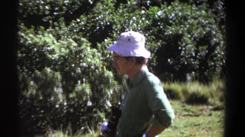 The man remains in profile in the 1969 Kenya home movie, passing bright foliage while carrying camera equipment at his side.