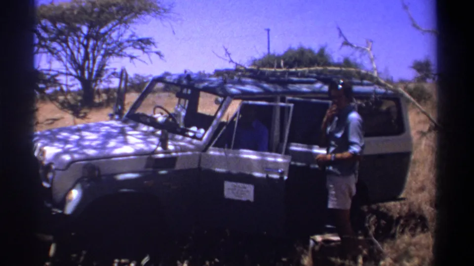 1969 Kenya 8mm frame showing an adult man beside a rugged safari vehicle in dry terrain.