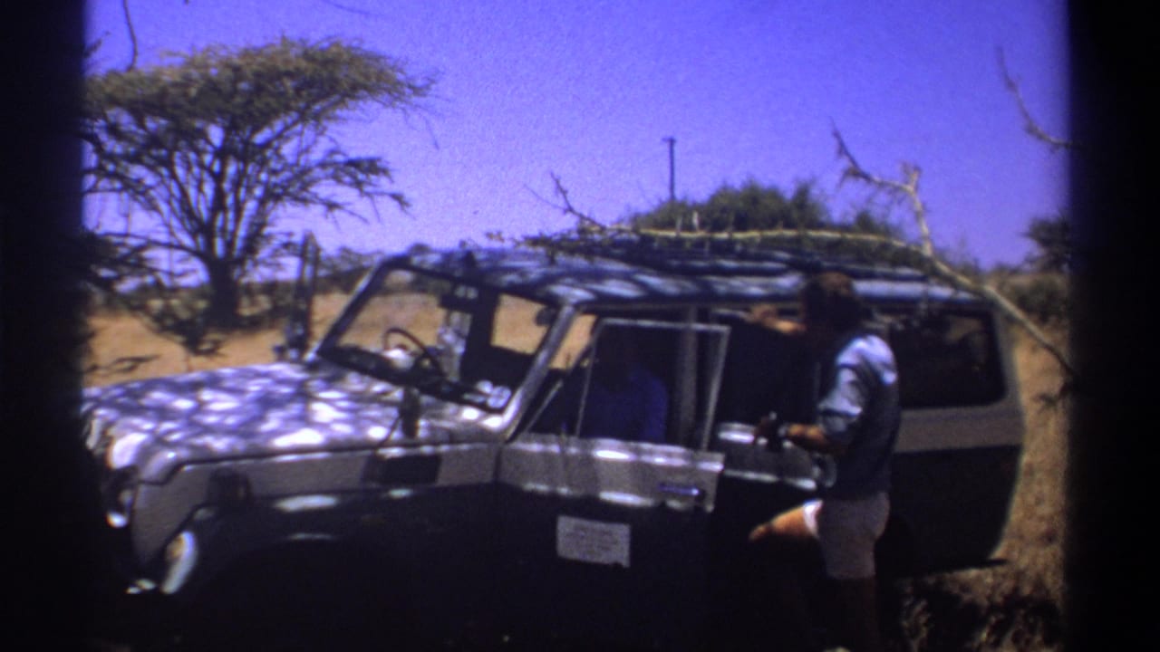1969 Kenya home-movie frame showing an adult man beside an open dark safari vehicle under bright sun.