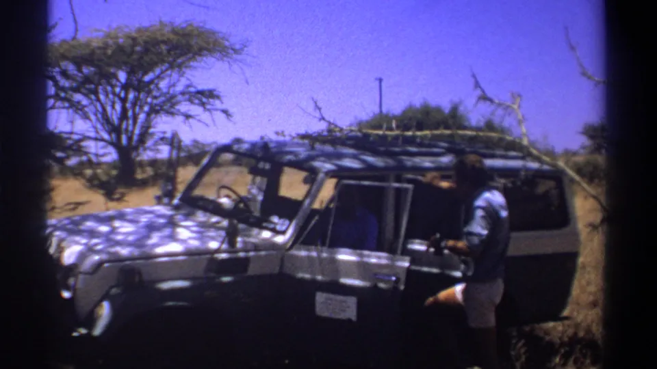 1969 Kenya home-movie frame showing an adult man beside an open dark safari vehicle under bright sun.