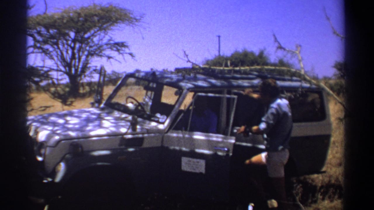 Man stands near the open passenger door of a roof-racked vehicle in savanna terrain.