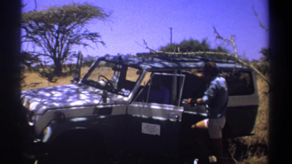 1969 Kenya frame with a man in shorts near an open safari vehicle door and roof rack loaded with branches.