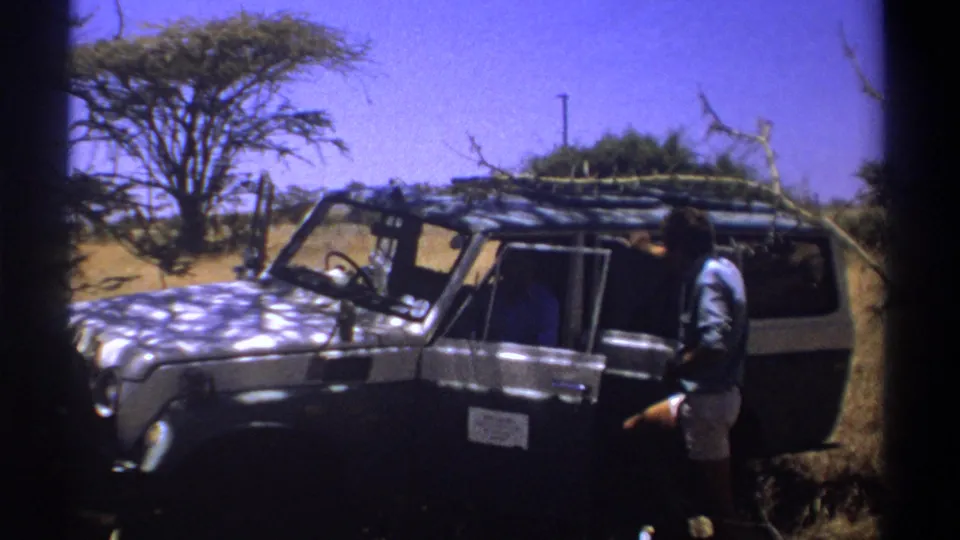 1969 Kenya home-movie still of a man by a dark safari vehicle, with dry grass and acacia-like trees visible.