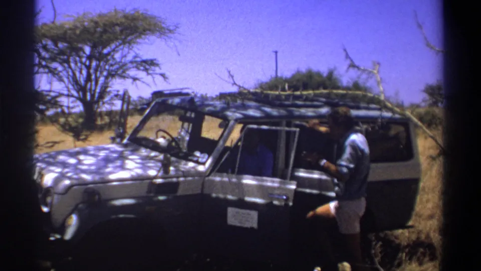 1969 Kenya frame showing a traveler beside a safari-style vehicle with branches on the roof rack.