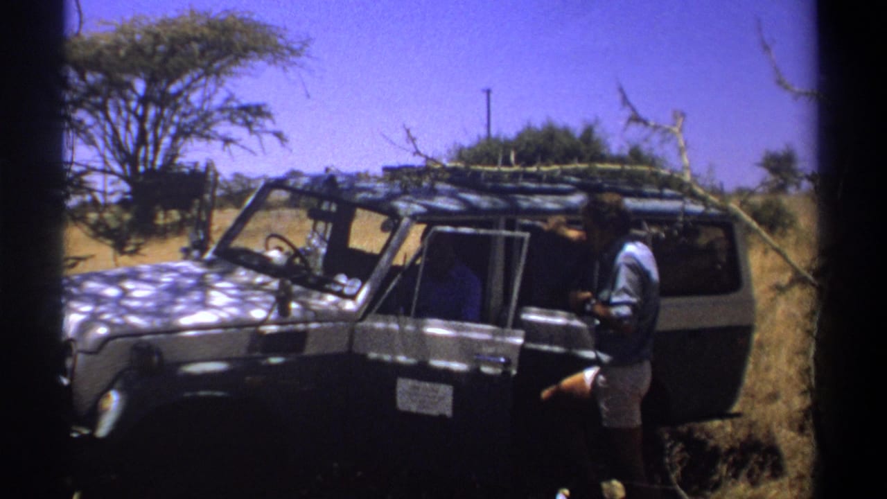 1969 Kenya home-movie frame showing a man beside a dark safari vehicle with roof branches in dry savanna.