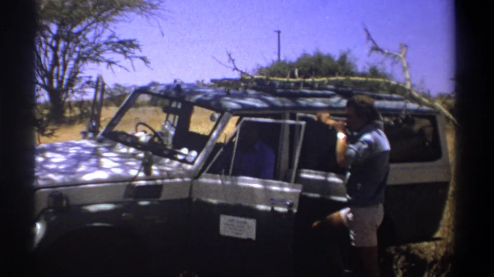 1969 Kenya home-movie frame showing a man gesturing or drinking beside a parked safari vehicle.