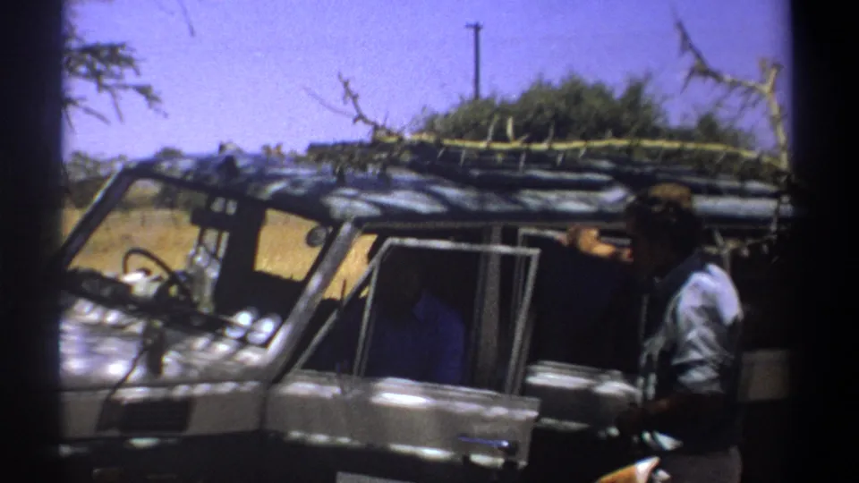 1969 Kenya frame with closer handheld view of the safari vehicle, roof branches, and man at right edge.