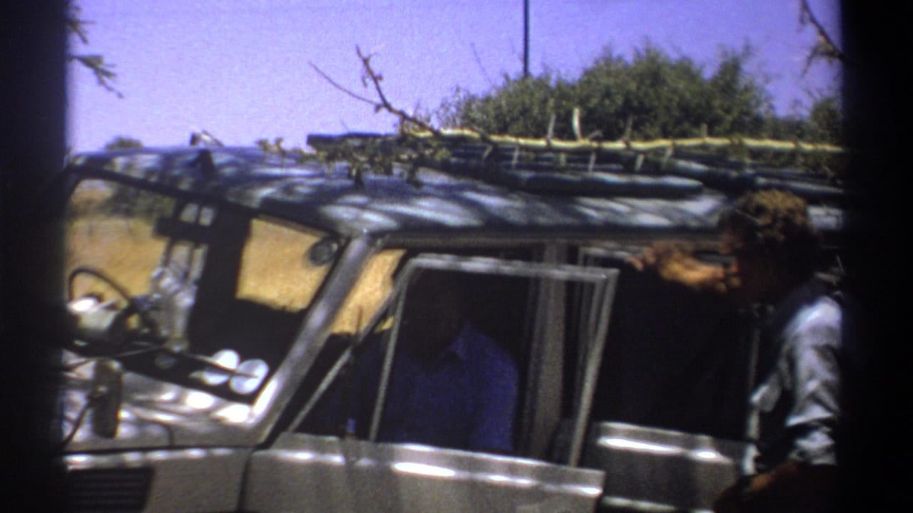 1969 Kenya 8mm frame showing two adult men by a rugged vehicle loaded with branches or gear on the roof.