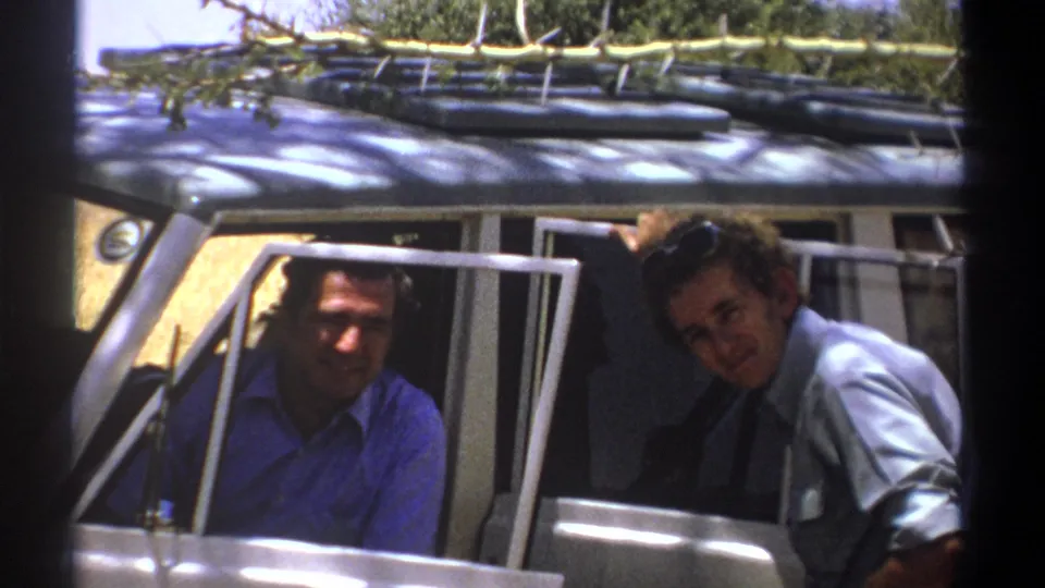 1969 Kenya 8mm frame with two adult men smiling near a rugged vehicle carrying tied gear or branches on its roof.