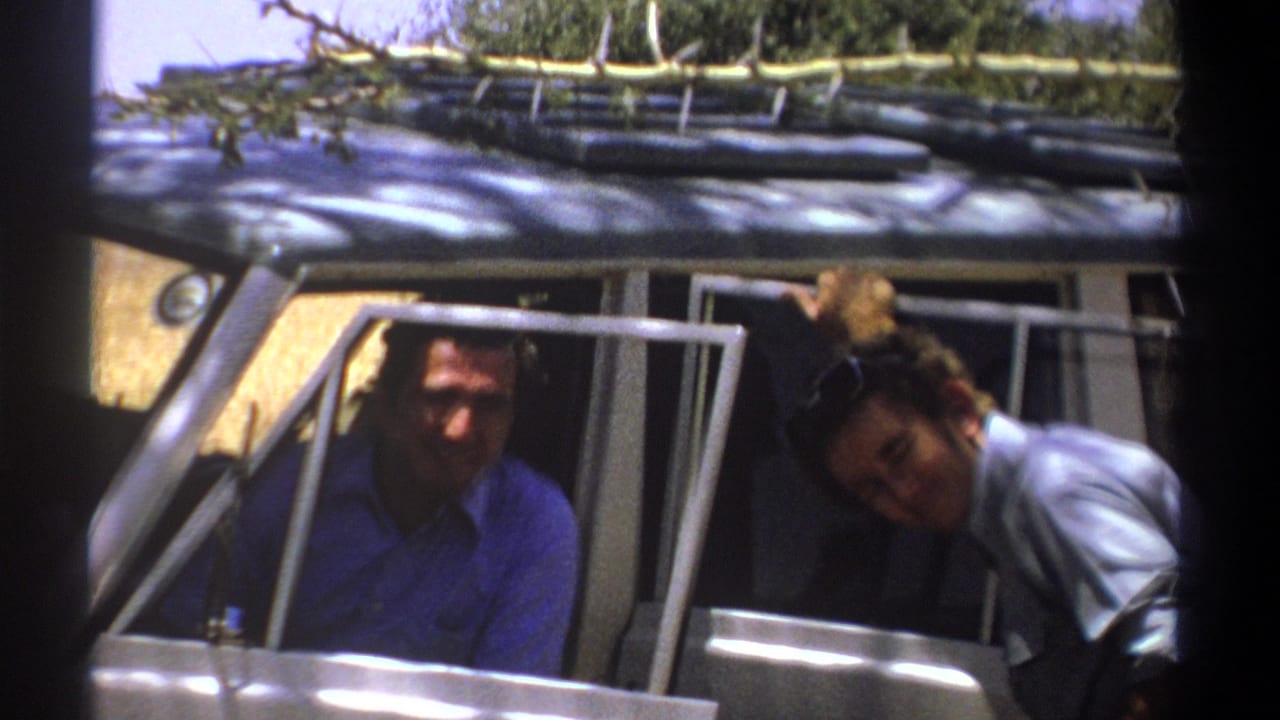 1969 Kenya home-movie frame showing two adult men smiling near open doors of a classic vehicle with foliage on the roof rack.