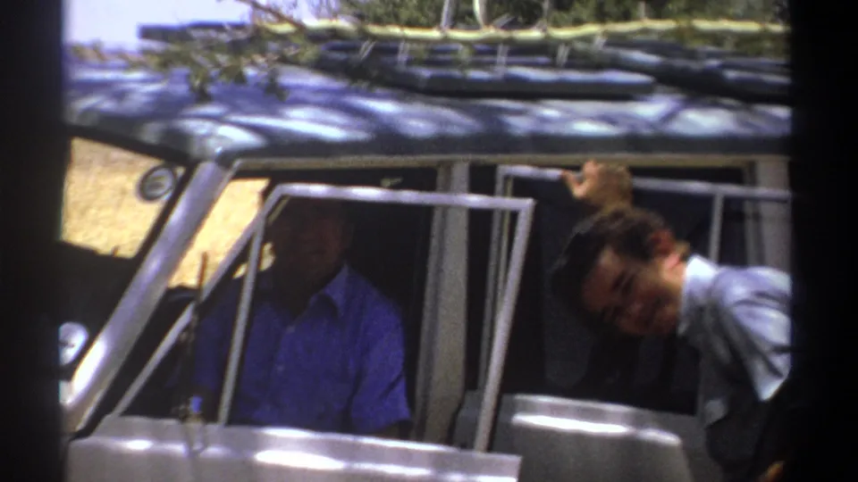1969 Kenya home-movie frame of an open vehicle, with one man shadowed inside and another leaning beside the door.