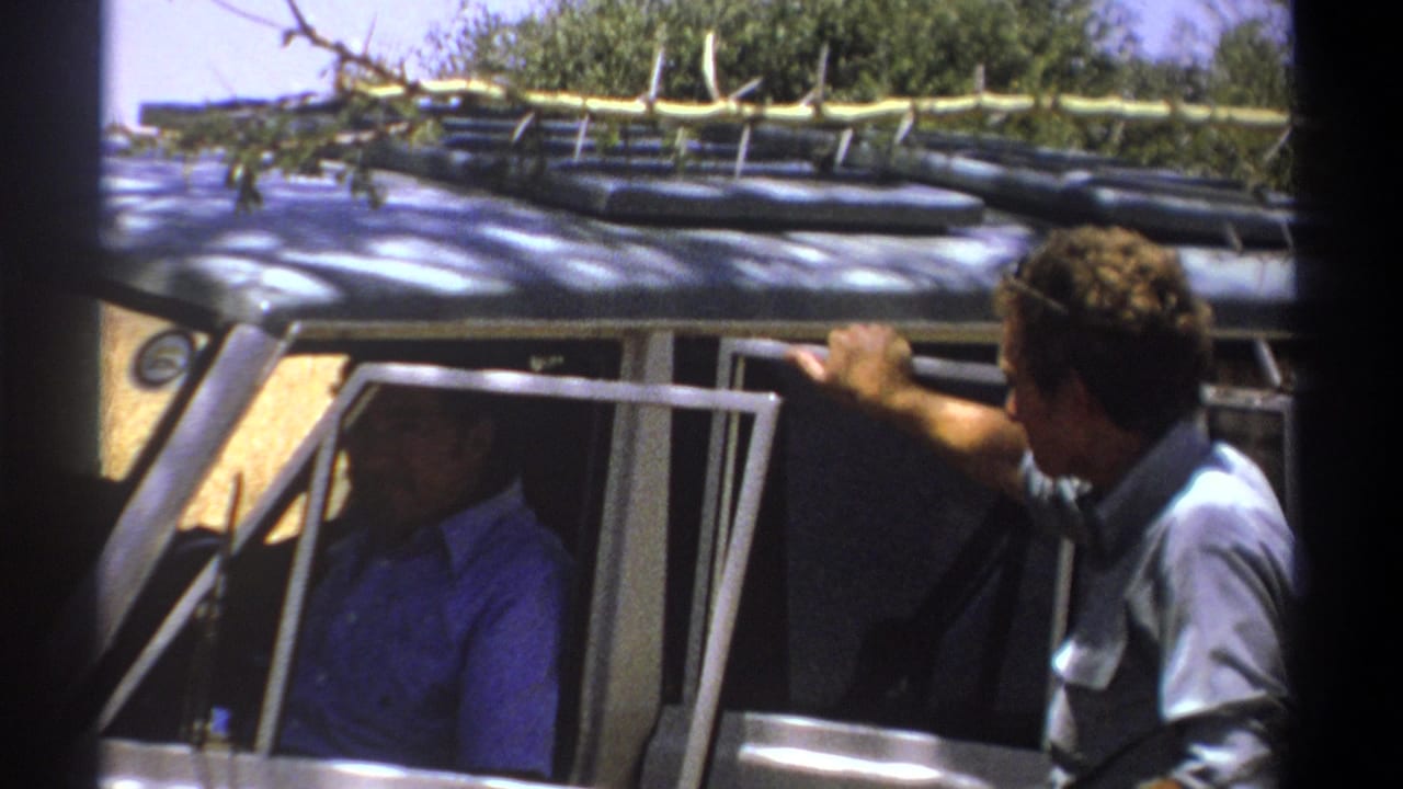1969 Kenya home-movie frame of two adult men at an open safari vehicle, with a roof rack and branch visible above.