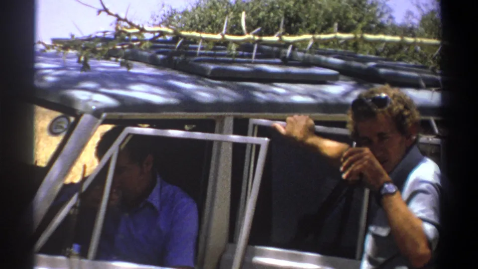 1969 Kenya frame showing a man with sunglasses on his head beside an open safari vehicle, with another man partly visible inside.