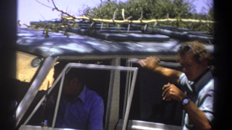1969 Kenya home-movie frame of two adult men at a safari vehicle, one seated inside and one leaning near the window.
