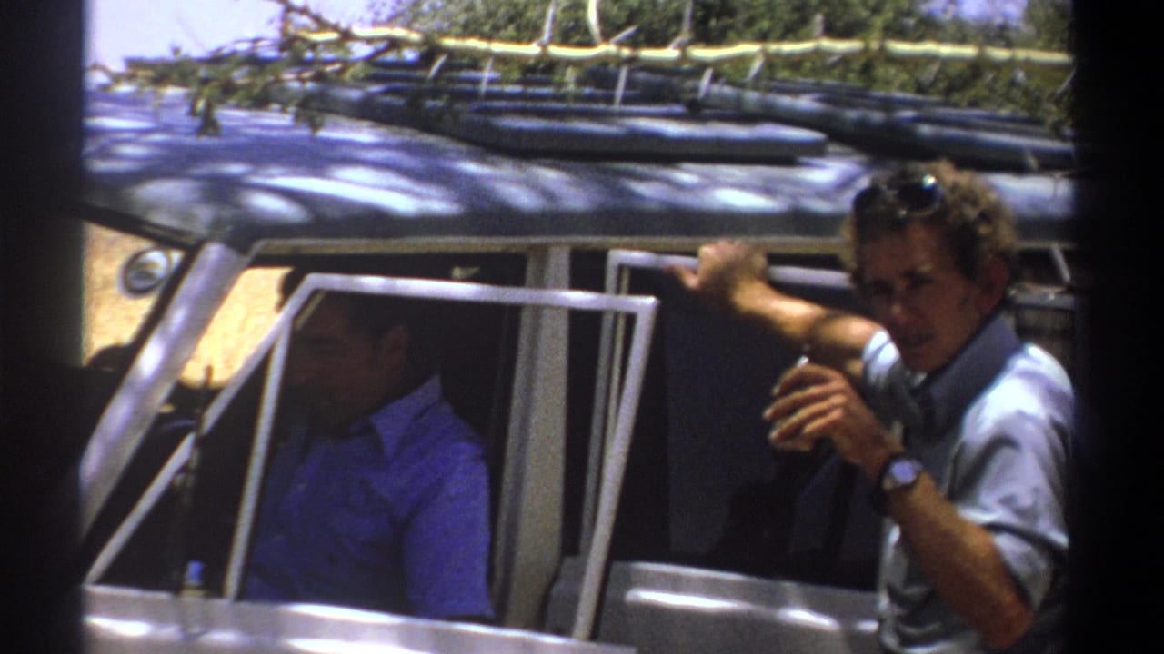 Man at a vehicle window gestures toward the camera while another man sits inside.