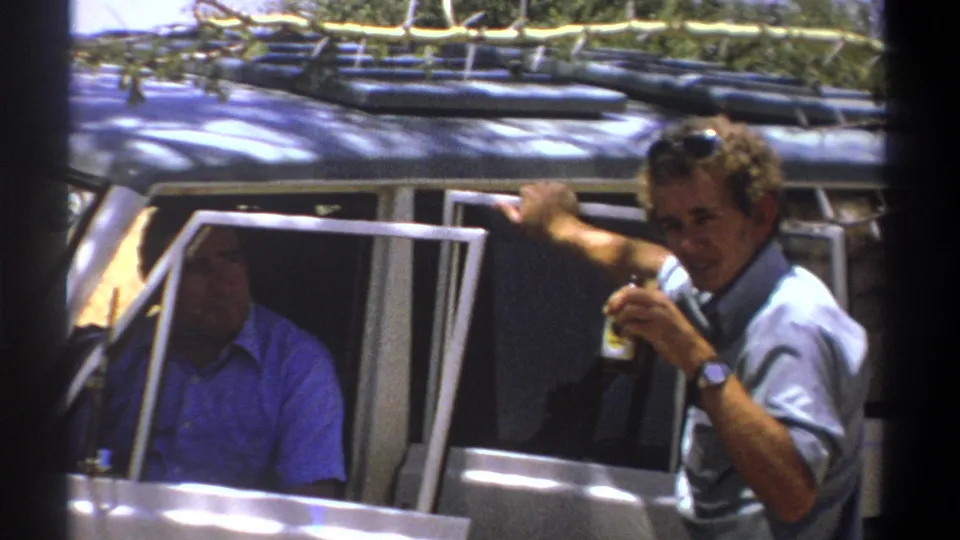 A 1969 Kenya home-movie frame shows a man holding a small object beside a roof-racked vehicle.