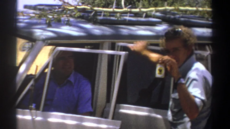 1969 Kenya safari footage frame with a man beside a vehicle raising a small badge or tag while a passenger is visible inside.