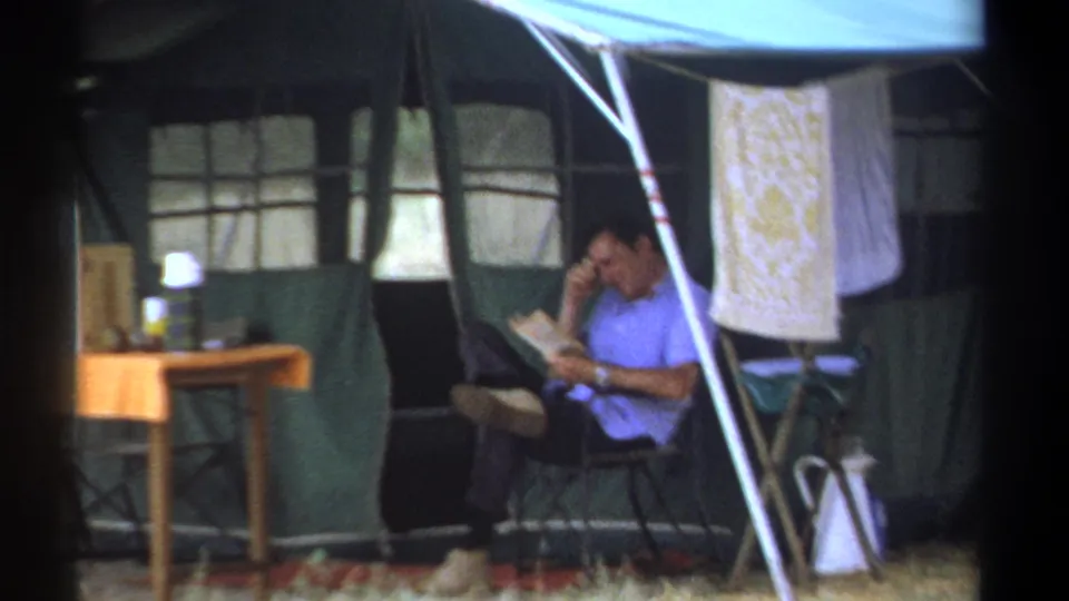 1969 Kenya camping scene with a man seated beside a canvas tent, folding furniture, a table, and hanging cloth.