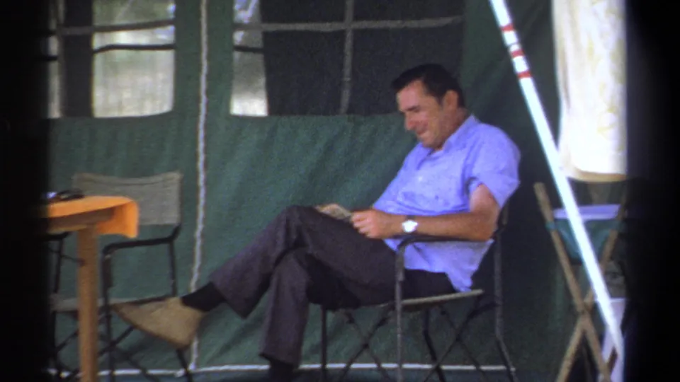 In 1969 Kenya footage, an older man sits in a folding chair inside a green canvas tent while reading a newspaper.