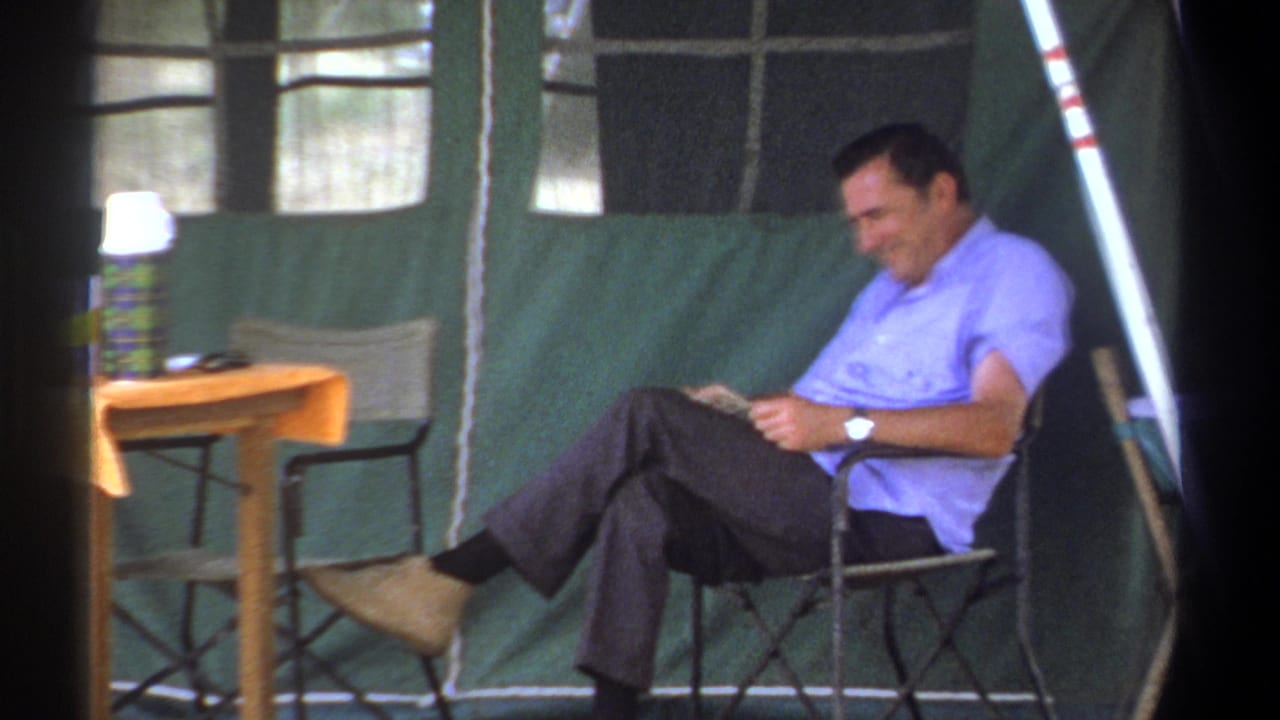 The man remains seated in the Kenya tent, reading near a small wooden table with a thermos visible.