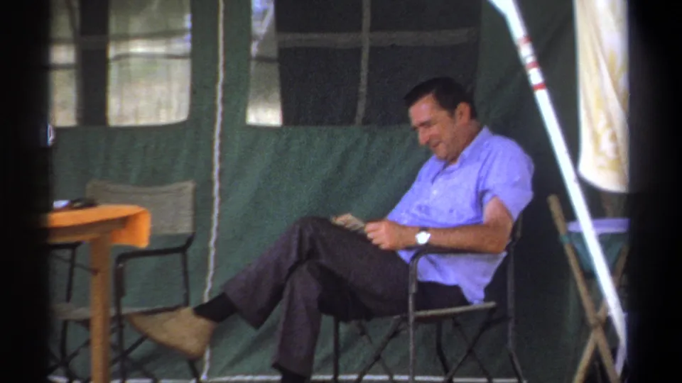 A home-movie frame shows the older man in a light blue shirt reading with legs crossed in the Kenya camp tent.