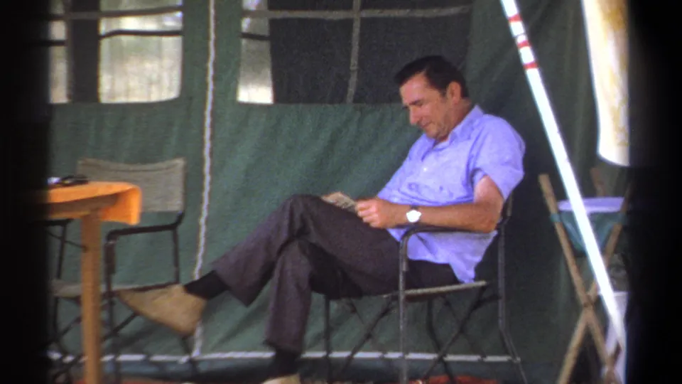 At the end of the clip, the seated older man continues reading inside the green tent, with table and camp gear nearby.