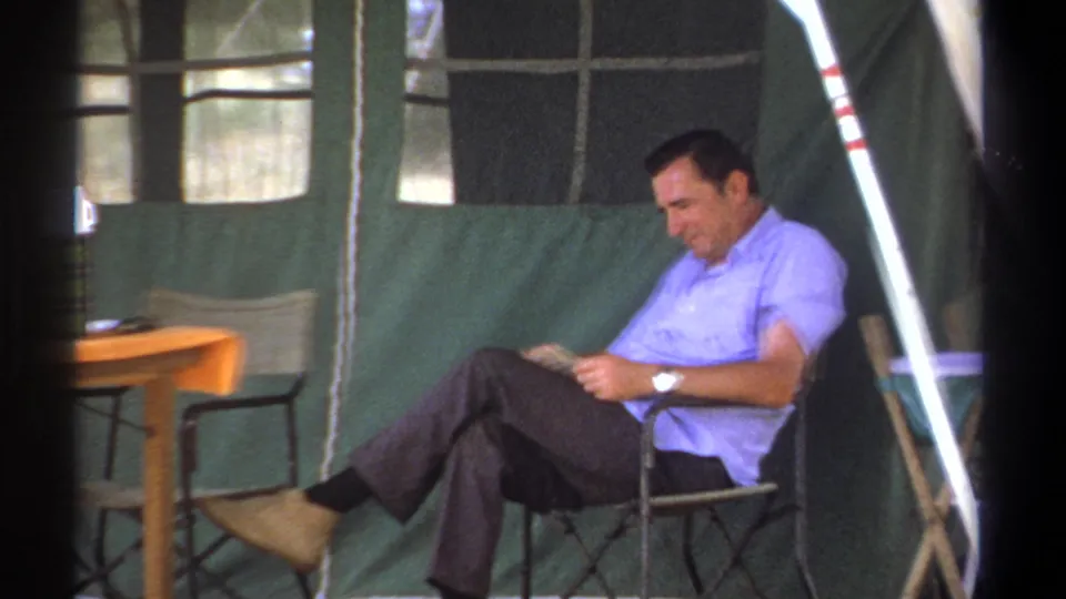 In Kenya, 1969, a man in a light blue shirt reads while seated inside a green canvas tent.