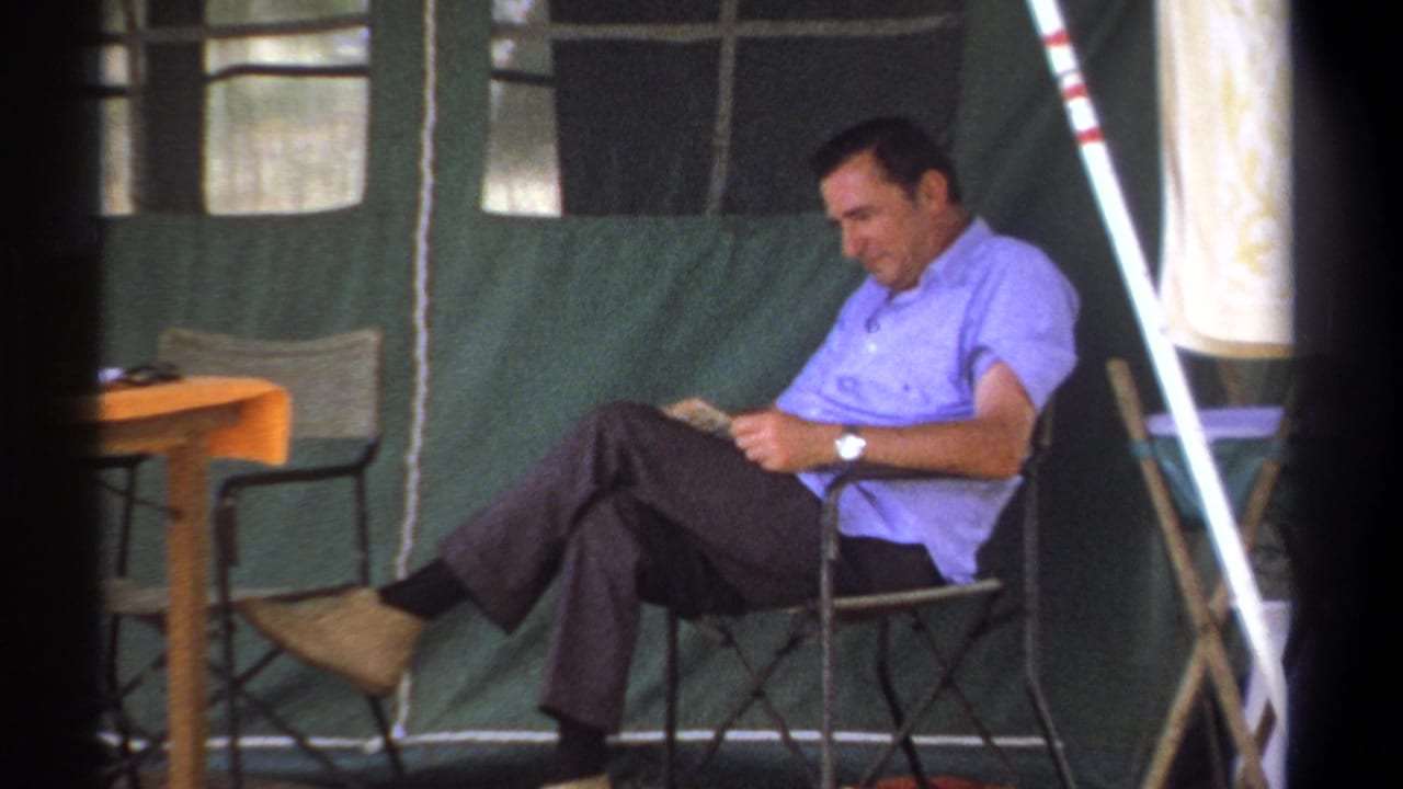 Man reading in a chair beside a small table inside a tent.