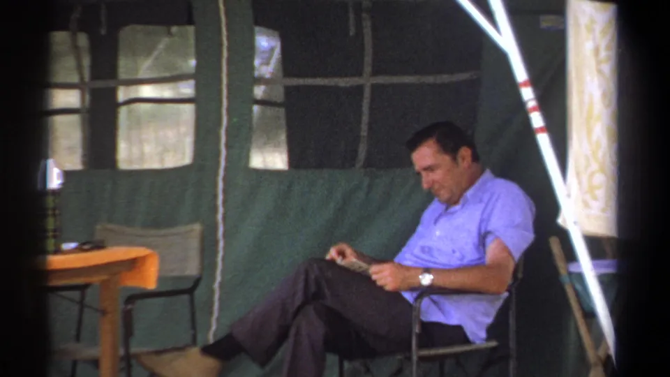 Inside a green canvas tent in Kenya, 1969, a man lowers his gaze toward reading material in his lap.