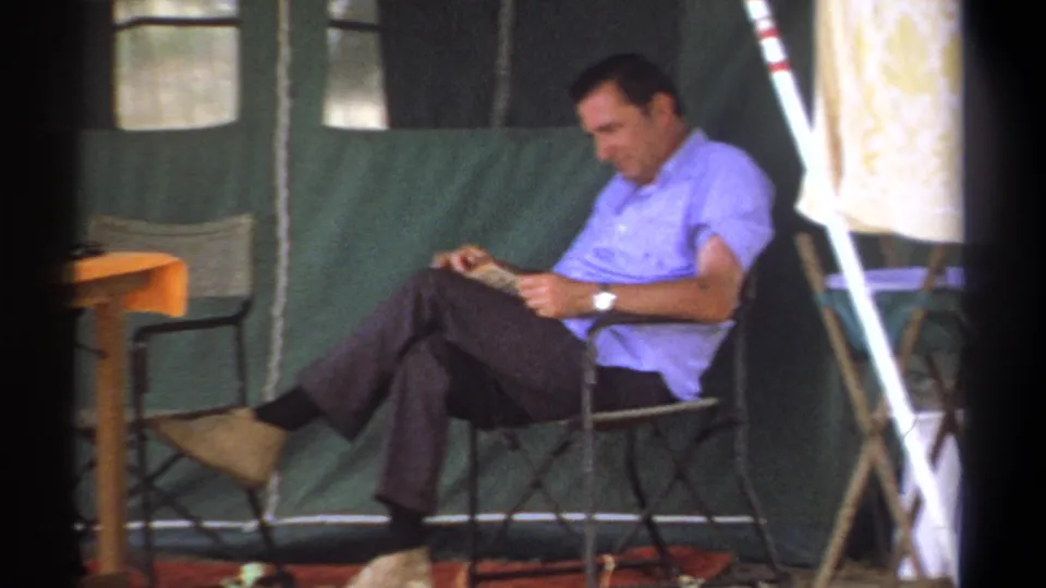 A man in a light blue shirt holds a book while resting inside a 1969 Kenya camping tent.