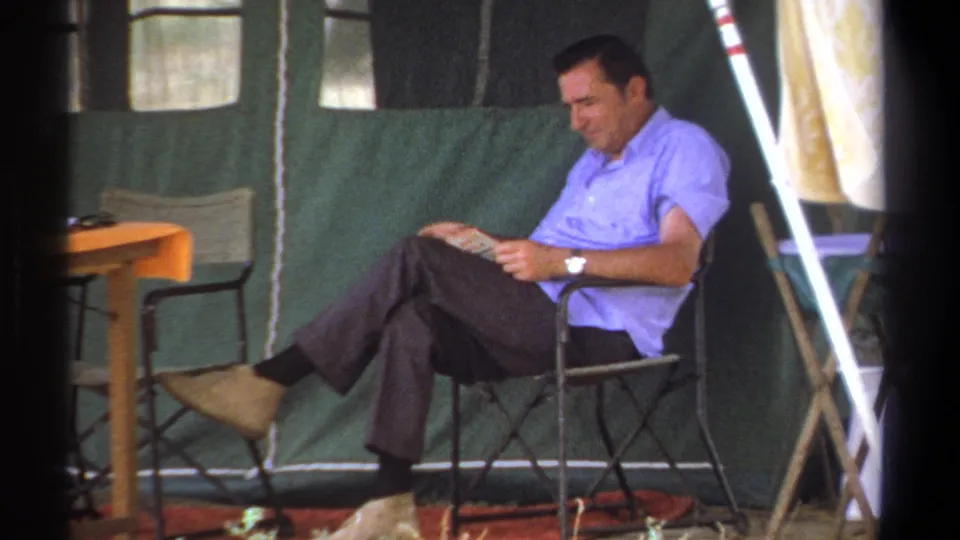In Kenya home-movie footage from 1969, a man in a light blue shirt reads while seated inside a green canvas tent.