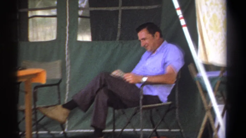 The 1969 Kenya home movie shows a closer view of the man reading in a folding chair against green canvas tent walls.