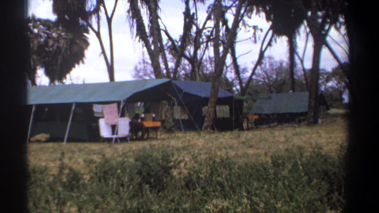 1969 Kenya home-movie frame showing canvas safari tents, chairs, a small table, dry grass, and tall trees around a campsite.