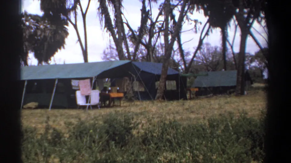 1969 Kenya home-movie frame showing canvas safari tents, chairs, a small table, dry grass, and tall trees around a campsite.