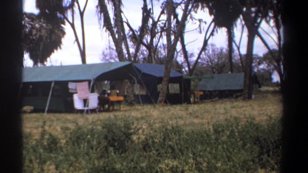 1969 Kenya campsite view with canvas tents, camp furniture, hanging cloth, and a distant person partly visible near the central tent.