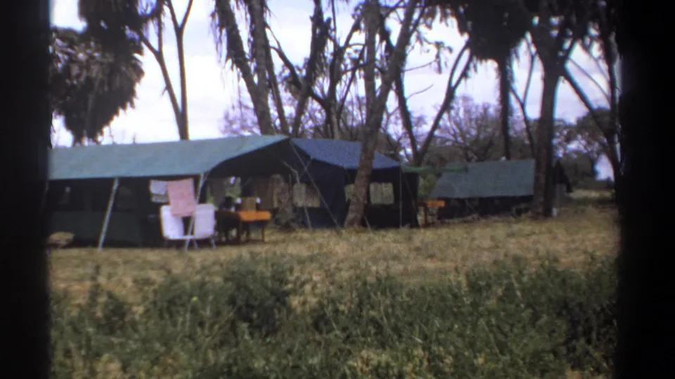 1969 Kenya campsite view with canvas tents, camp furniture, hanging cloth, and a distant person partly visible near the central tent.